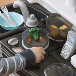 Kids playing with a realistic pretend play kitchen set featuring toy fridge, oven, and cookware